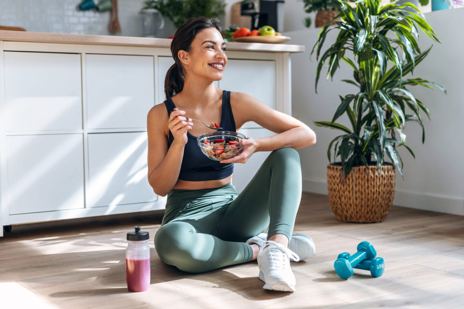 Shot of athletic woman eating a healthy bowl of muesli with fruit sitting on floor in the kitchen at home